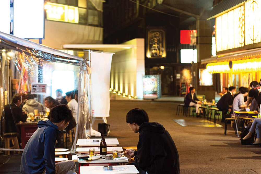 Outdoor night scene at a street food market in Malaysia, with diners seated at tables under ambient lighting, enjoying meals