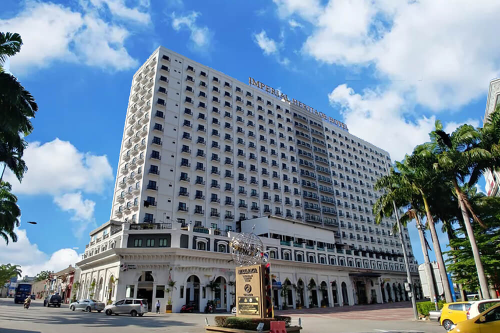 Large, white multi-story hotel with entrances at the base under a clear blue sky, palm trees, and a bright yellow car